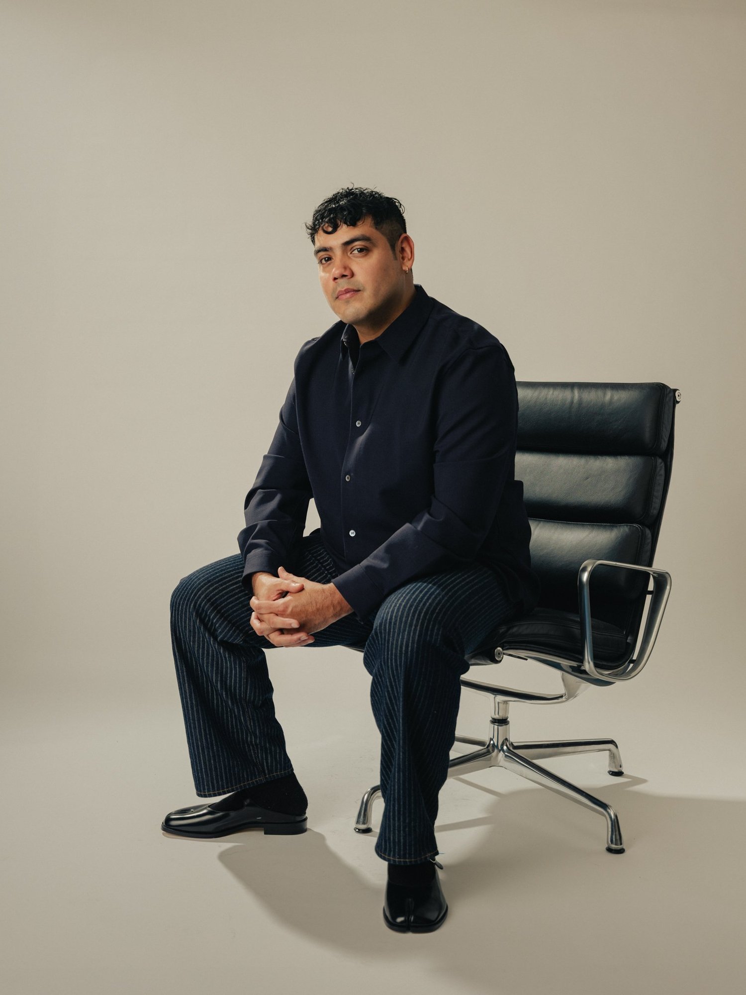 Saul Flores seated in a studio, wearing a black shirt and trousers